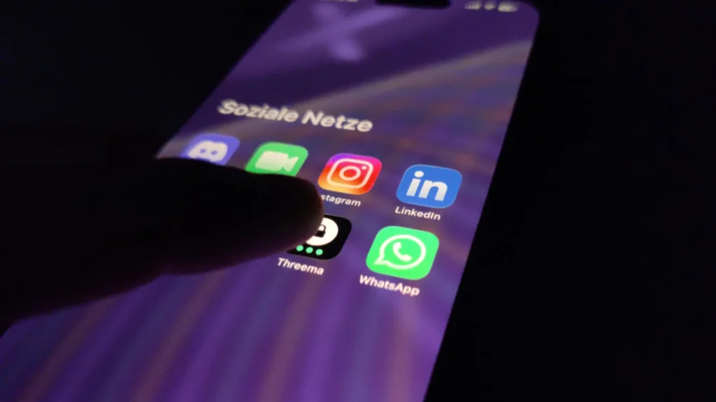 Finger tippt im Dunkeln auf einen Smartphone-Ordner mit WhatsApp, Threema und LinkedIn: Symbolbild zur EU-Chatkontrolle 2026